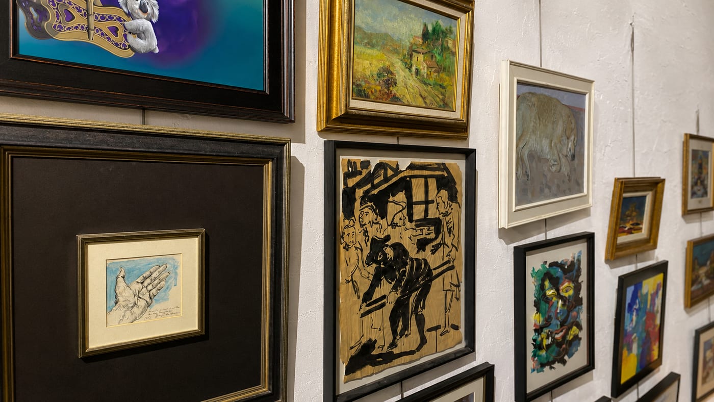 A gallery wall seen at an angle, hung with framed works of many kinds — an ink drawing, a Tuscan landscape in a gilt frame, a study of a hand, small bright abstracts
