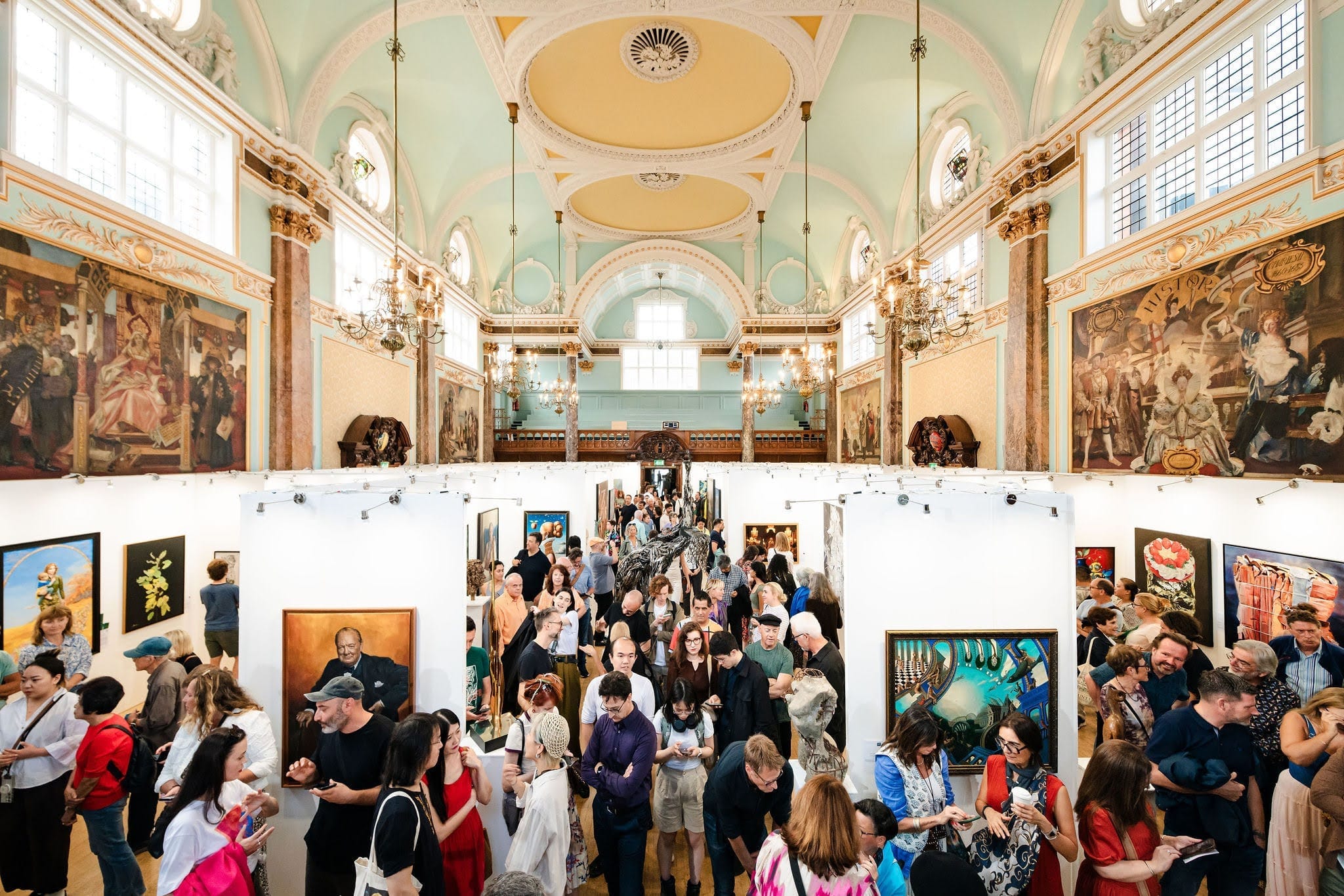The main hall of Chelsea Old Town Hall at the opening of the 2025 London Art Biennale, packed with visitors among the paintings, chandeliers hanging from the vaulted ceiling